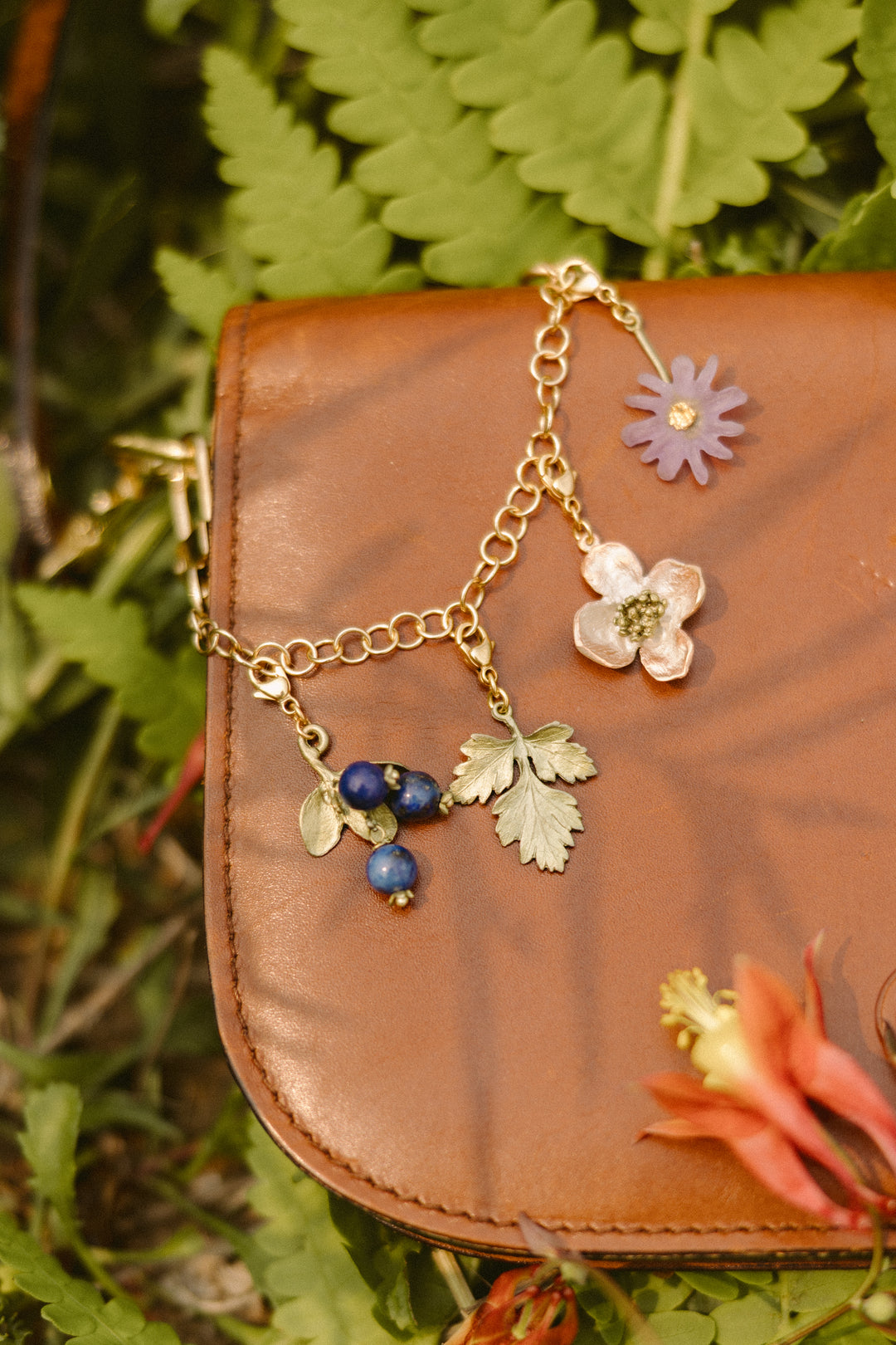 A brown leather bag on green ferns, decorated with a gold chain featuring flower and leaf charms in purple, pink, green, and blue.