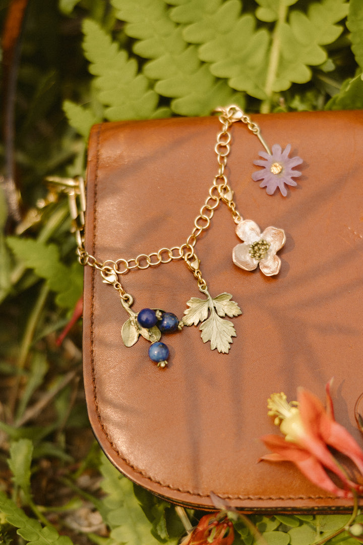 A brown leather bag on green ferns, decorated with a gold chain featuring flower and leaf charms in purple, pink, green, and blue.