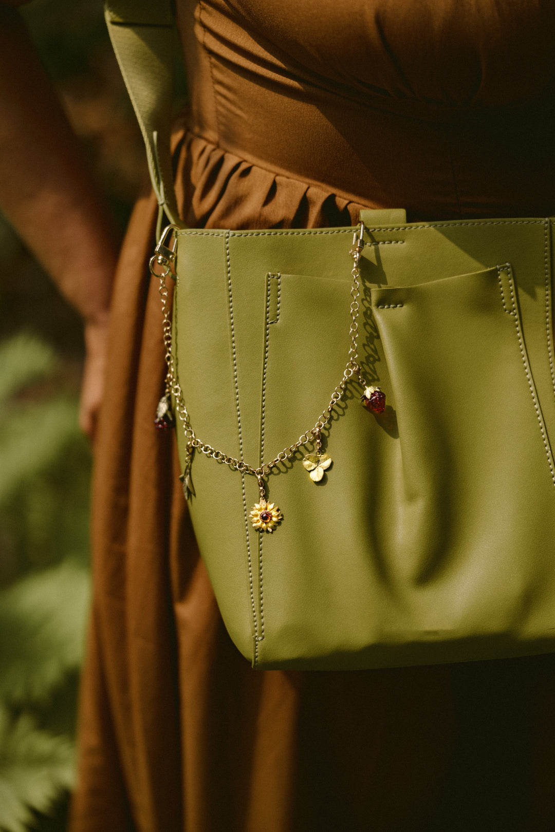 A person in a brown dress carries a green handbag decorated with a chain and small charms, including a flower and a butterfly.