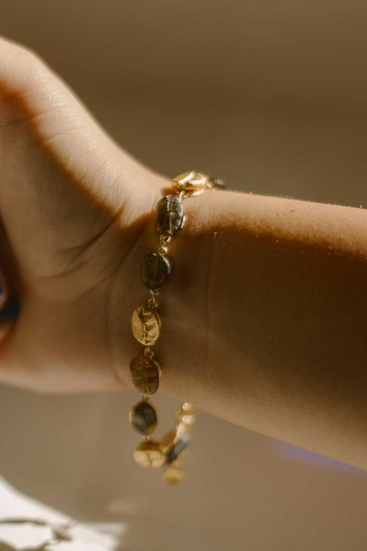 A close-up of a wrist wearing the Roasted Coffee Bean Bracelet, crafted in gold-plated bronze with coffee beans, captured in soft natural light.