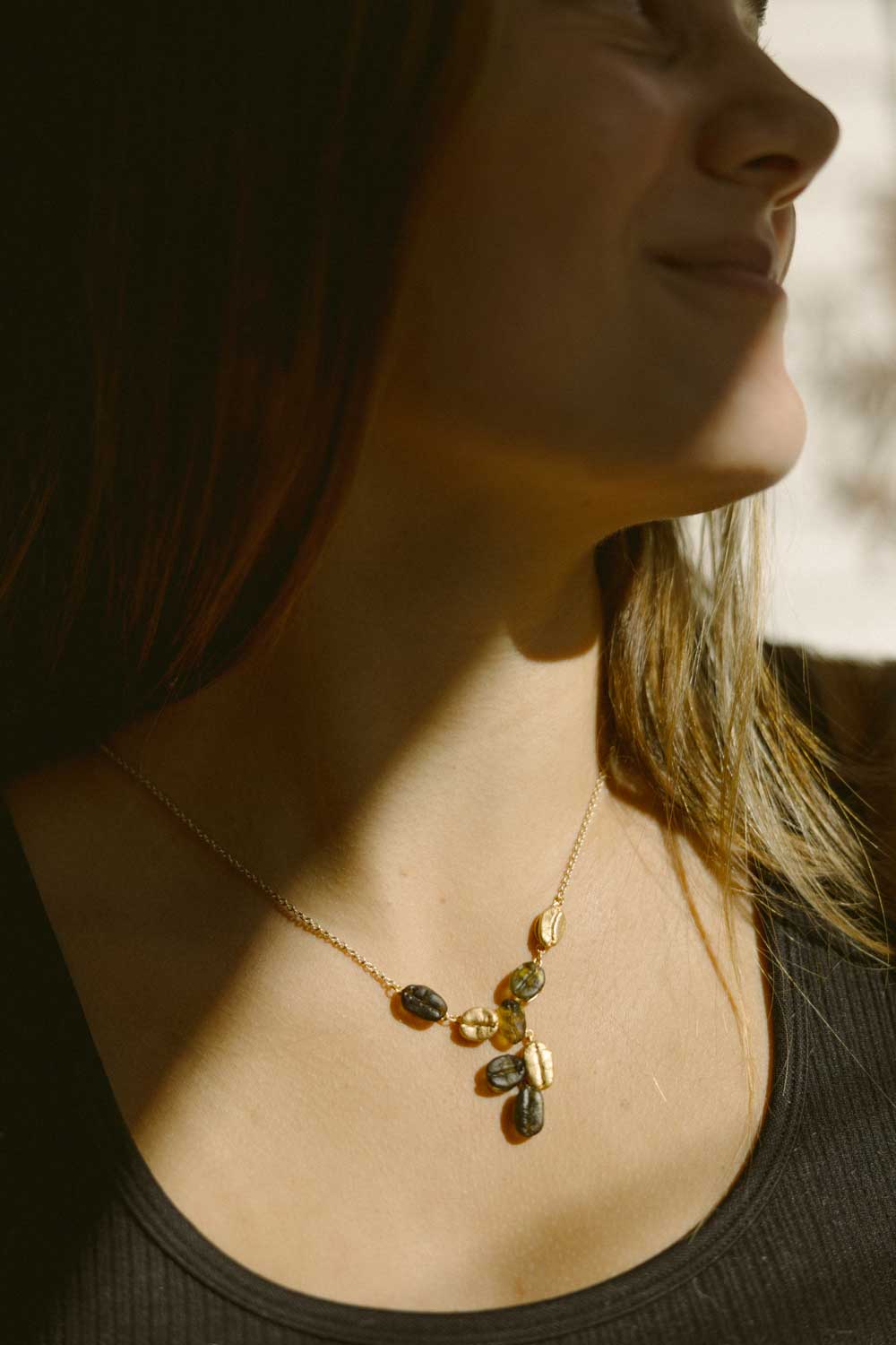 A person wears a sleeveless black top and the Roasted Coffee Bean Pendant necklace, shown from the shoulders up with sunlight casting shadows on their face.