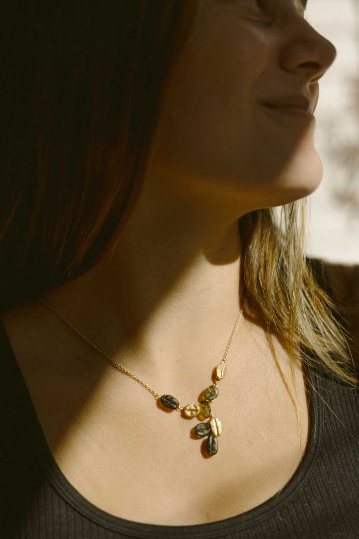 A person wears a sleeveless black top and the Roasted Coffee Bean Pendant necklace, shown from the shoulders up with sunlight casting shadows on their face.