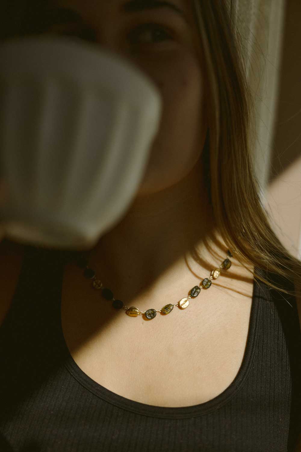 A woman in a black tank top wears the Roasted Coffee Bean Necklace, holding a white cup near her face, partially obscuring her mouth as warm sunlight shines across her chest and neck.