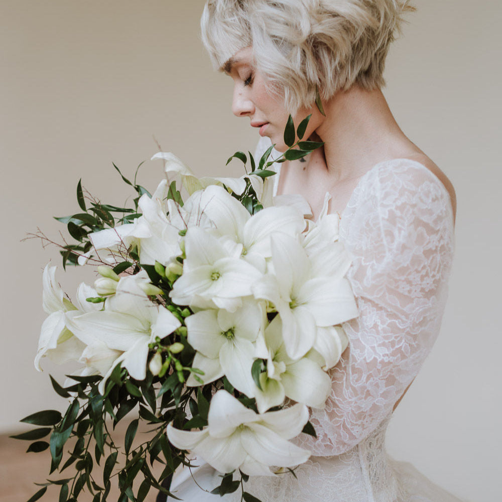 A woman in a lace wedding dress holds a large bouquet of white lilies and greenery, looking down with a neutral expression against a plain background.