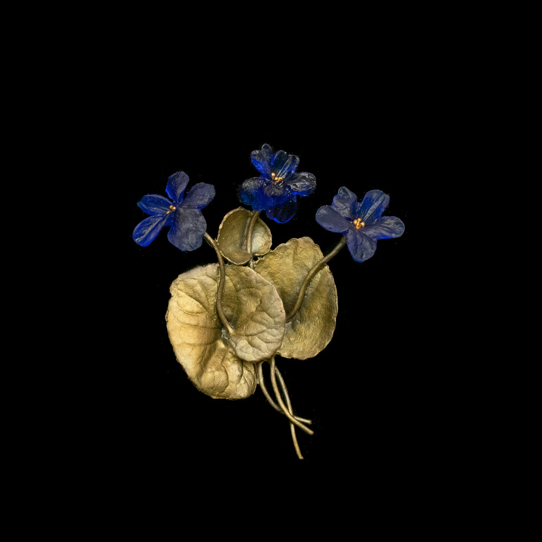 A brooch featuring three blue flowers with yellow centers and three textured golden leaves, all set against a black background. The stems and leaves are metallic with a slightly curved design.