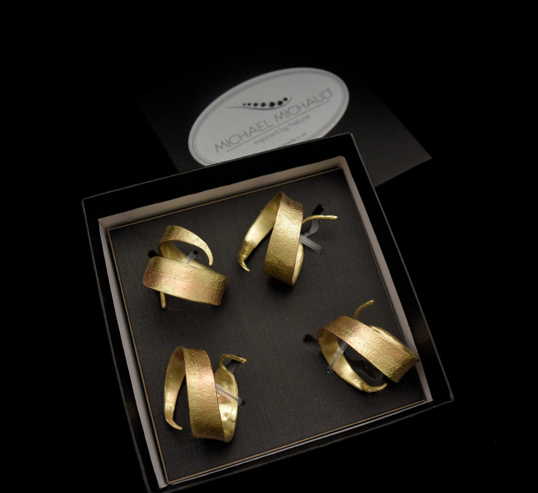 A set of four gold-toned, textured earrings of varying abstract shapes displayed in a black box with a Michael Michaud card in the background, all set against a black backdrop—perfect for fans of elegant home accessories.