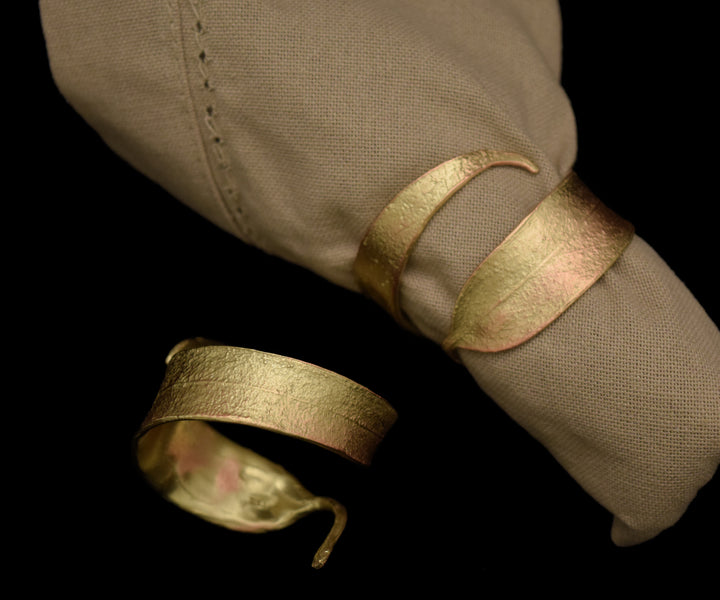 Two simple, textured gold napkin rings are shown—one encircling a folded beige cloth napkin, the other resting beside it on a black background—an elegant home accessory.