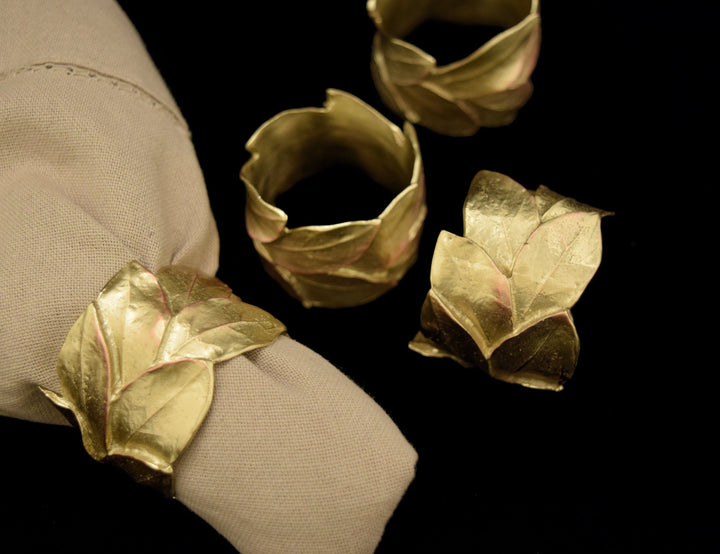 four golden napkin rings in leaf design, one is placed around a beige fabric napkin