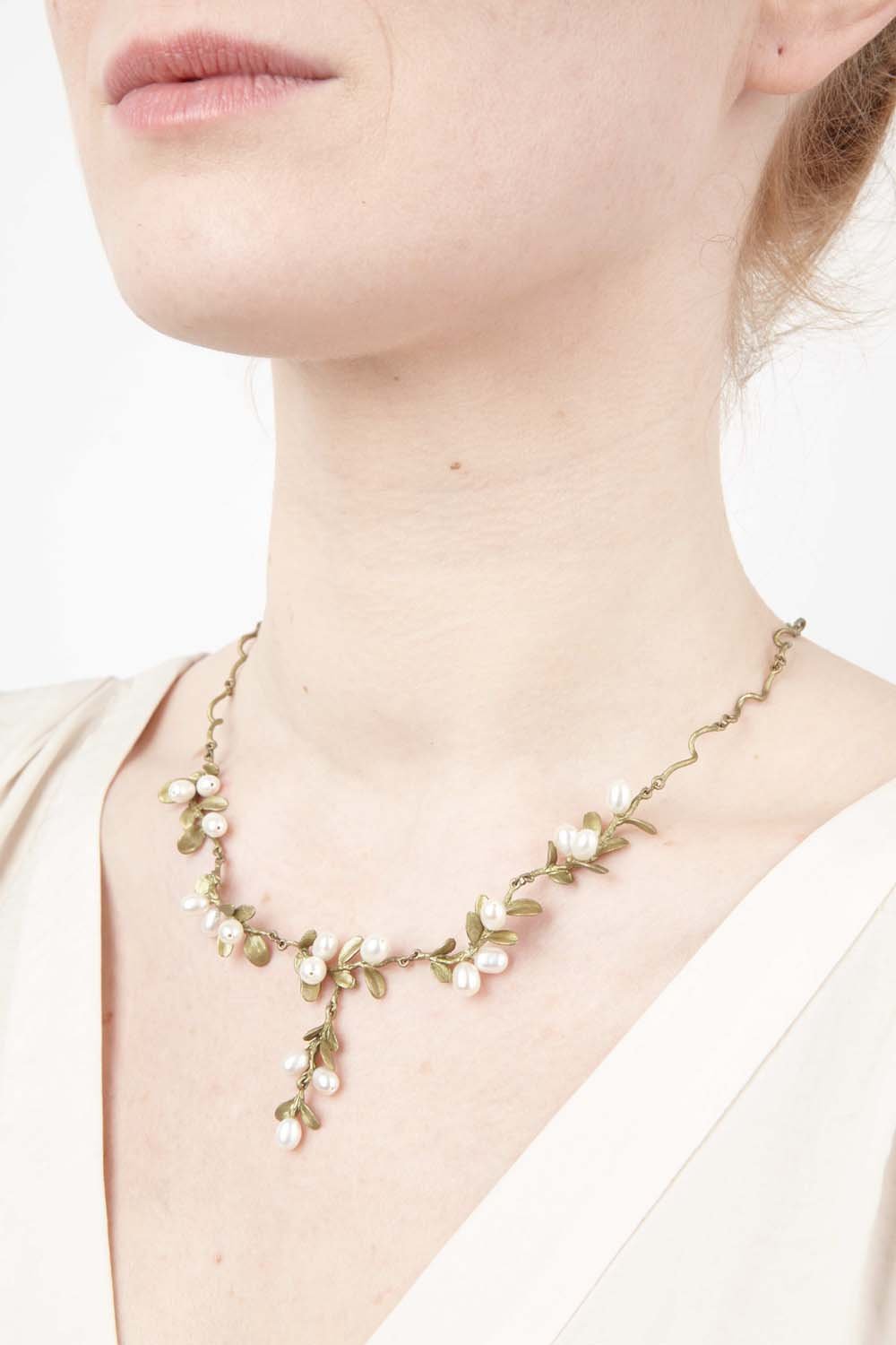 A close-up of a woman wearing a cream top and a delicate Mount Vernon jewelry necklace featuring small white and pink flowers with hand patinated bronze green leaves on a gold chain, displayed against a plain white background.