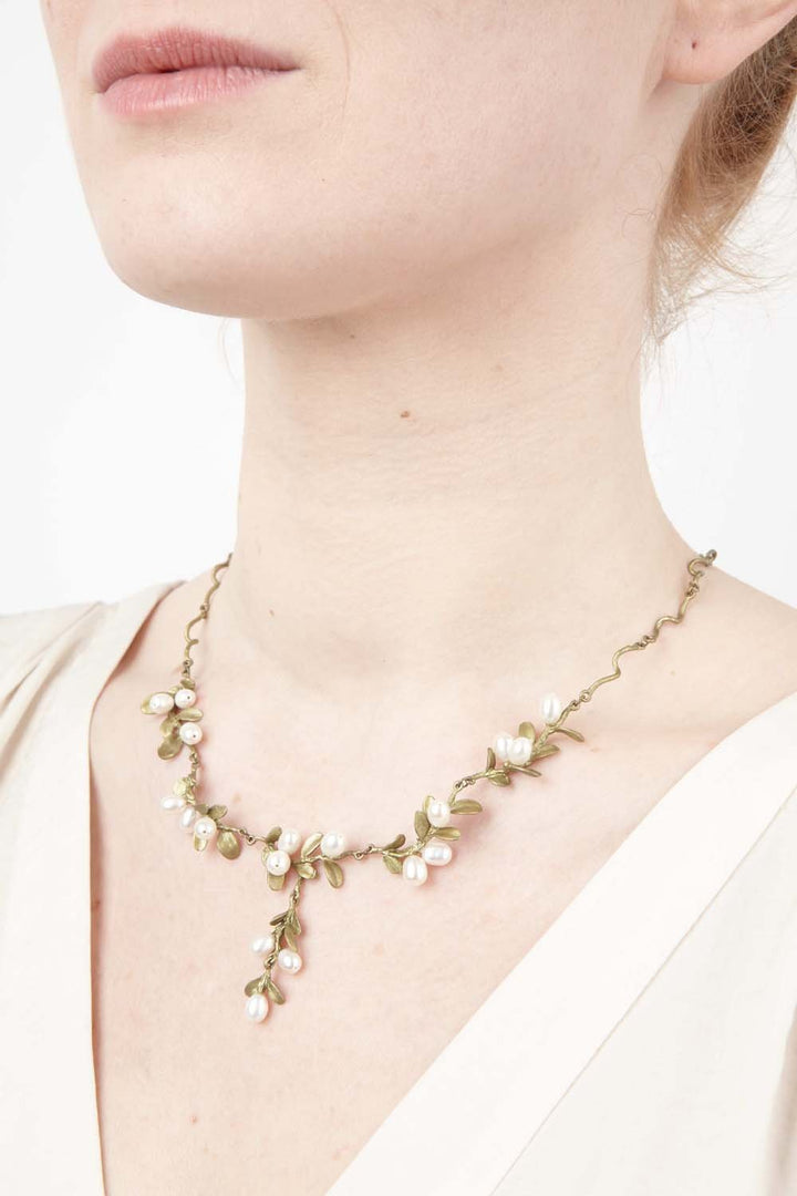 A close-up of a woman wearing a cream top and a delicate Mount Vernon jewelry necklace featuring small white and pink flowers with hand patinated bronze green leaves on a gold chain, displayed against a plain white background.