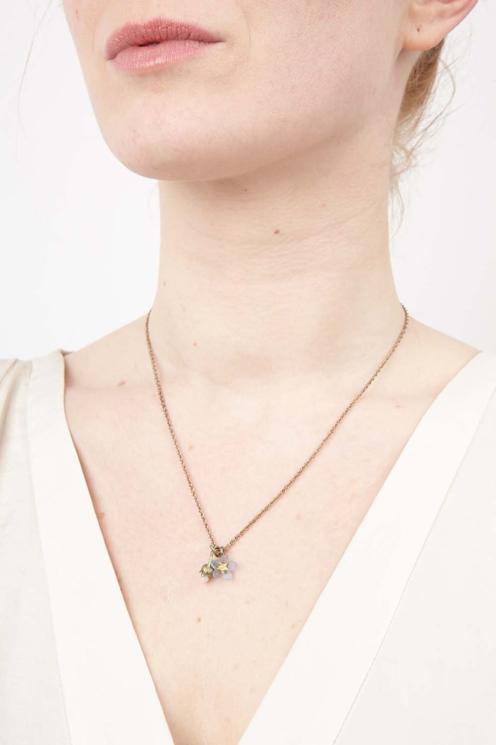 A woman wears a cream V-neck top and a delicate gold chain necklace featuring a hand patinated bronze Forget Me Not pendant. The image is cropped to show her neck, lower face, and upper chest, highlighting this elegant remembrance jewelry.