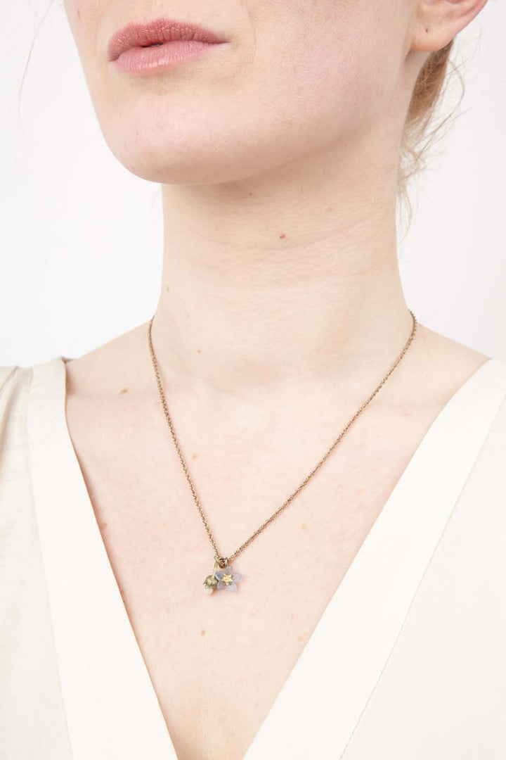 A woman wears a cream V-neck top and a delicate gold chain necklace featuring a hand patinated bronze Forget Me Not pendant. The image is cropped to show her neck, lower face, and upper chest, highlighting this elegant remembrance jewelry.