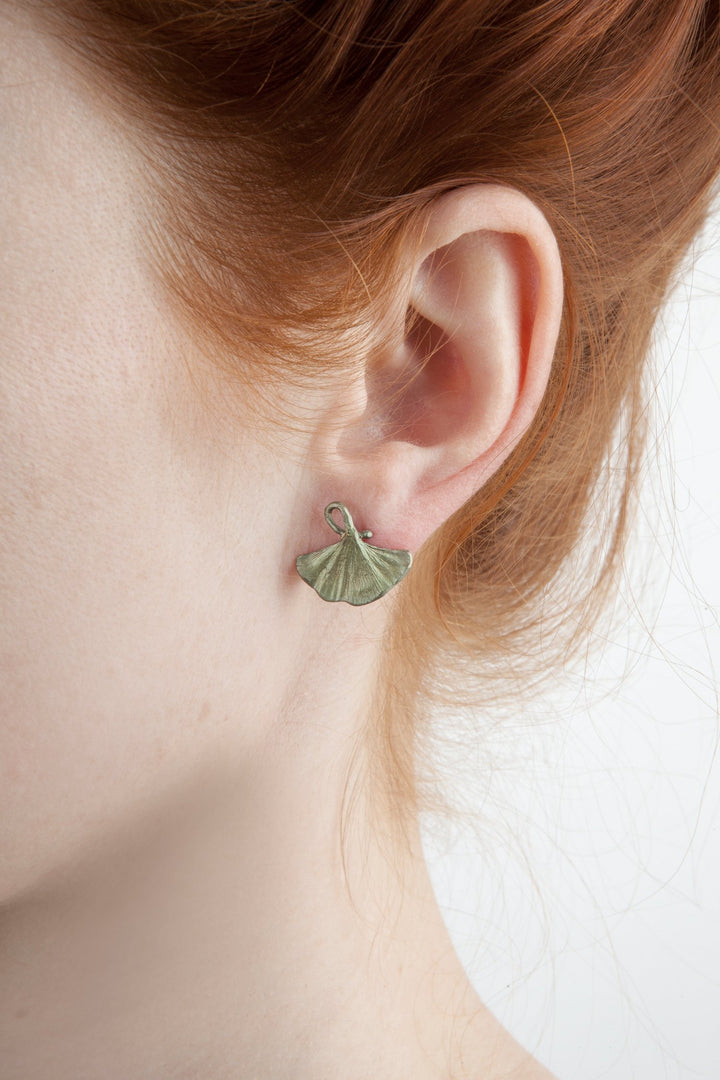 Green ginkgo leaf-shaped stud earrings on ear with red hair background.